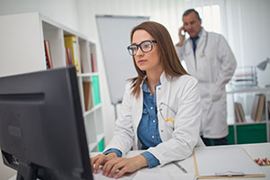 Woman doctor typing at computer with male doctor on phone in the background
